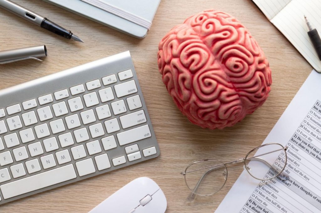 Uma foto de um cérebro realista visto de cima. O cérebro está em uma mesa rodeado de objetos como teclado, caneta, mouse, um óculos e uma folha de papel.