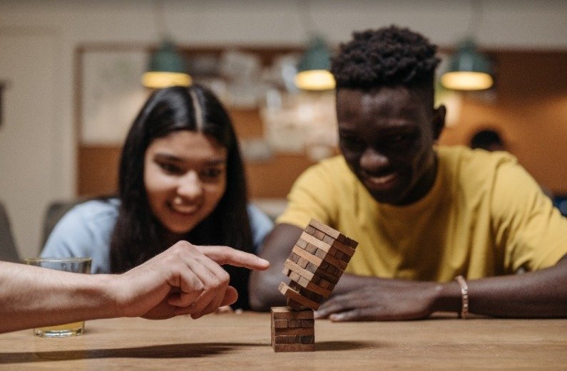 Dois amigos (fora de foco), uma mulher branca de cabelo liso escuro e um homem negro de cabelo curto observam com empolgação um dedo de uma pessoa branca derrubando uma torre do jogo "JENGA"