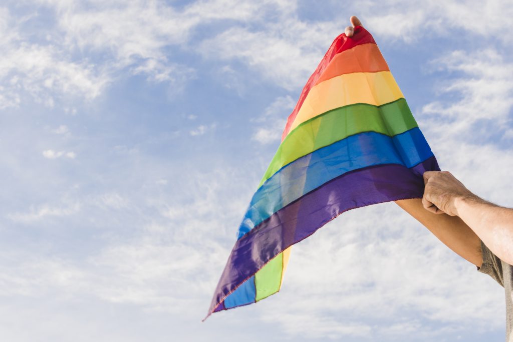 A imagem mostra os braços de um homem branco segurando uma grande bandeira com as cores da comunidade LGBTQIA+, e ao fundo há um céu azul.