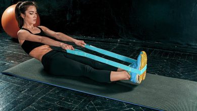 A imagem traz uma mulher se exercitando com um elástico azul, em que está deitada com as pernas esticadas para cima com a ajuda do elástico extensor.