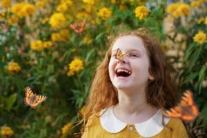A imagem retrata uma menina, com vestes amarelas, em um jardim repleto de flores e borboletas, sorrindo com uma borboleta pousada em seu nariz.