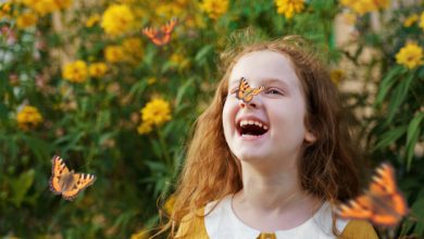 A imagem retrata uma menina, com vestes amarelas, em um jardim repleto de flores e borboletas, sorrindo com uma borboleta pousada em seu nariz.