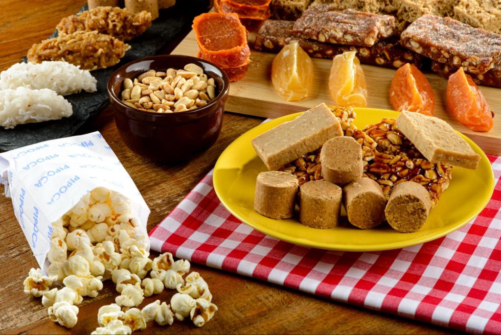 comidas de festa julina - paçoca, pipoca, amendoim, cocada e doce de batata doce e abóbora - em uma mesa com toalha quadriculada.