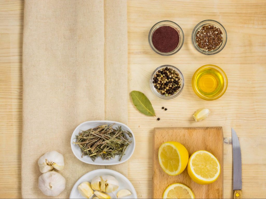 mesa com temperos - alecrim, alho, azeite, manjericão, pimenta...- e limão em uma mesa de madeira com um pano marrom.