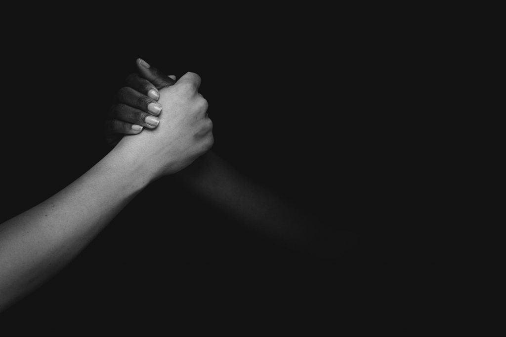 A imagem está em preto e branco e contém duas mãos dadas como laços de amizades. A mão posicionada na esquerda tem a pele branca e a da esquerda é negra.