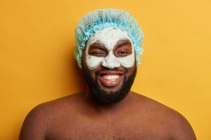 Homem negro, sorrindo com máscara facial aplicada no rosto, utilizando uma touca de banho azul com bolinhas brancas.
