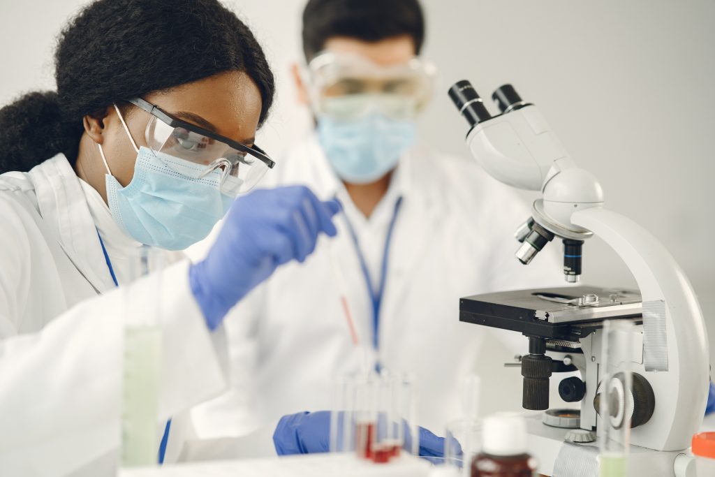 A imagem exibe uma mulher negra e um homem branco, ambos são profissionais da saúde, e estão em um laboratório analisando a coleta de exames médicos em um microscópio. 