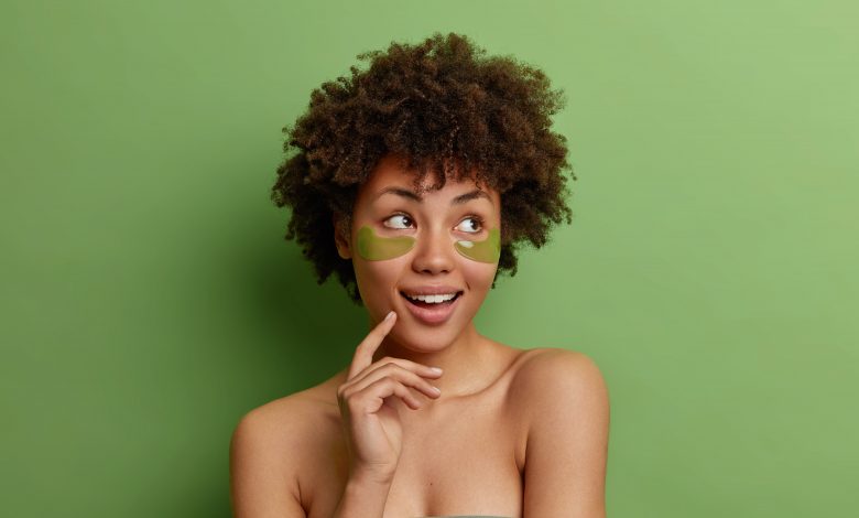 Mulher jovem negra, em um fundo verde, sorrindo com o dedo apoiado no rosto, enquanto realiza uma rotina de cuidado facial.
