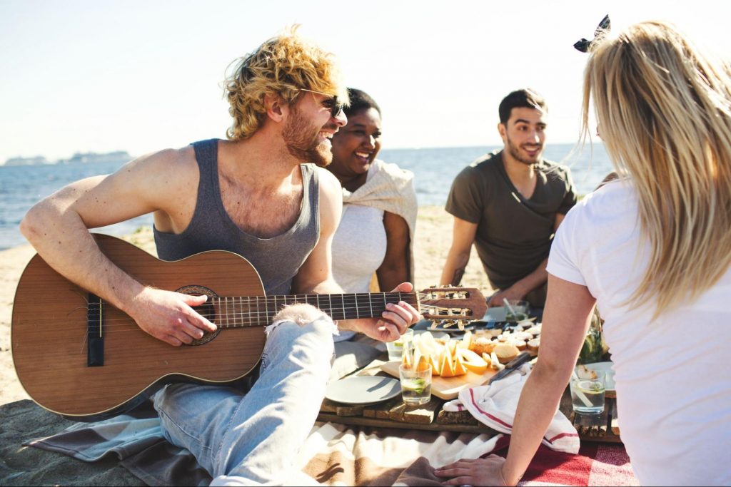 Roda de amigos sentados em um piquenique conversando e tocando música, selando amizades. A Imagem contém uma mulher branca loira de costas, um homem branco loiro tocando violão, uma mulher negra rindo e um homem branco conversando.