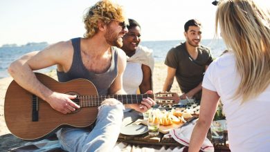 Pessoas sentadas em um piquenique conversando e tocando música, selando amizades. A Imagem contém uma mulher branca loira de costas, um homem branco loiro tocando violão, uma mulher negra rindo e um homem branco conversando.