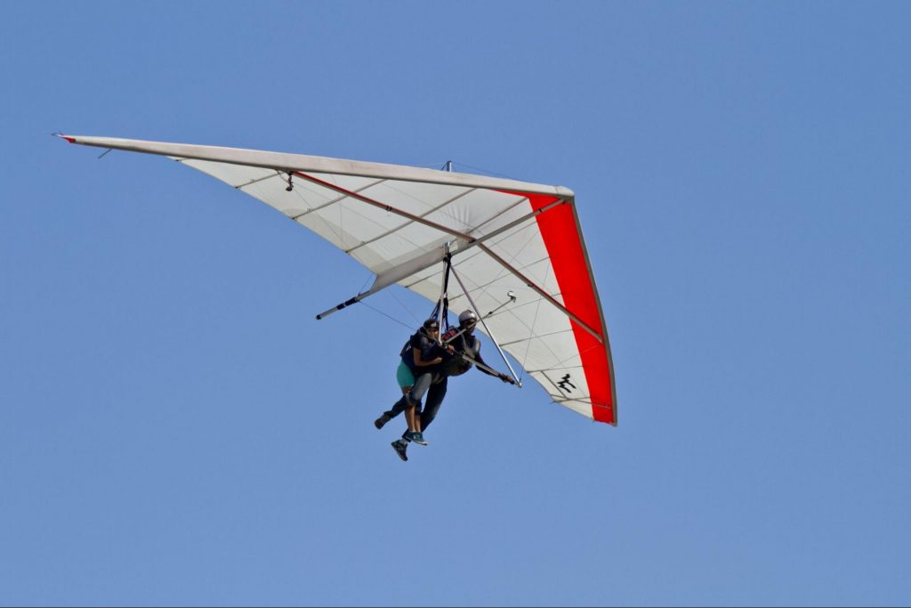 A imagem mostra a prática de esportes radicais, composta por duas pessoas realizando um voo de uma asa delta branca e vermelha. O fundo é composto apenas pelo céu completamente azul.