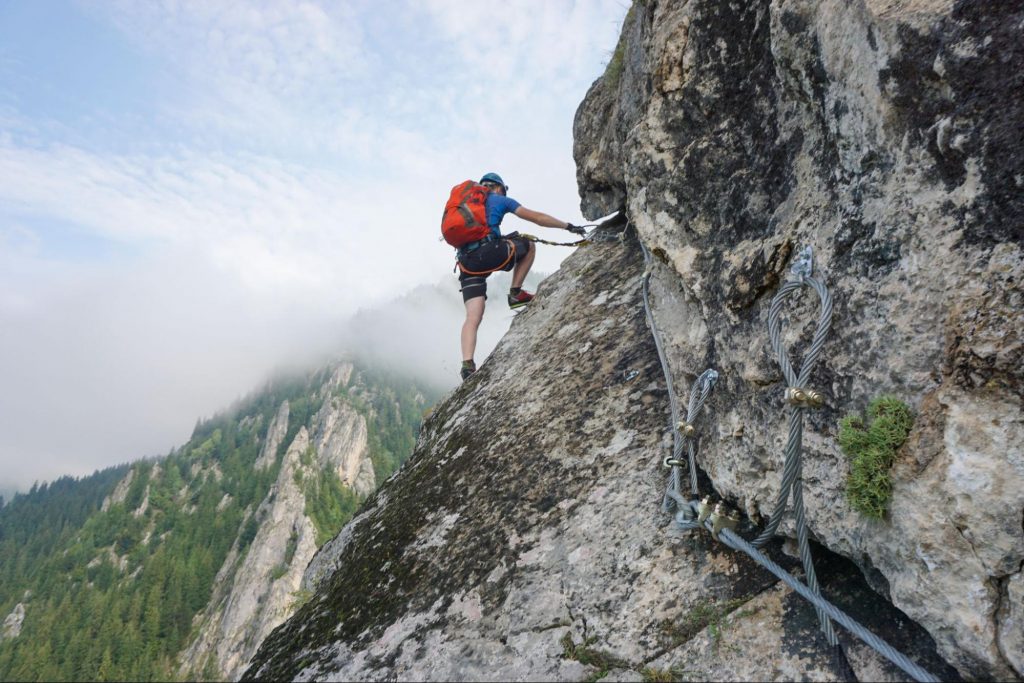 A imagem mostra a prática de esportes radicais, constituída por uma paisagem de uma montanha com um céu azul ao fundo. Ao centro, encontramos um homem escalando a parede da montanha.