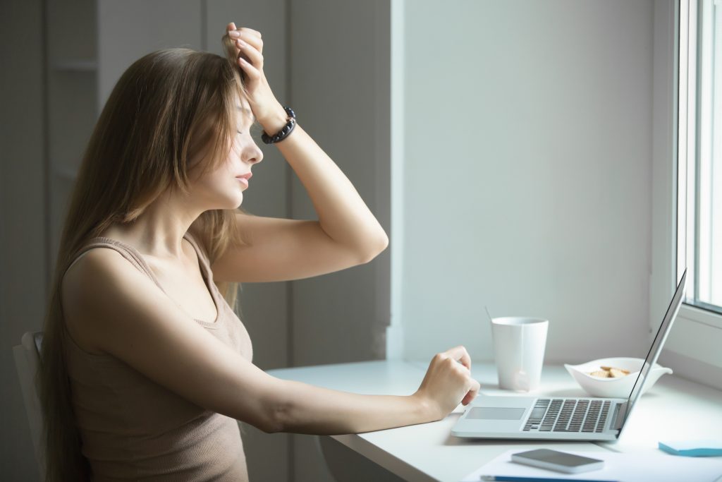 Imagem representando a ansiedade. Mulher branca sentada em frente ao computador em uma mesa branca, com a mão na cabeça, representando dor de cabeça.