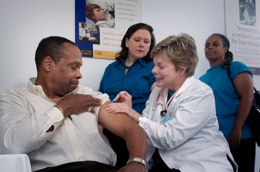 A imagem mostra um homem sendo vacinado por uma enfermeira,e duas mulheres observando no fundo