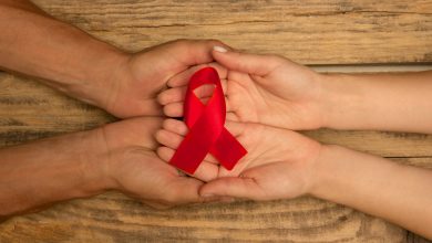 A imagem traz um laço vermelho, símbolo da conscientização do Dezembro Vermelho e do combate à pandemia da Aids, em mãos sobrepostas de duas pessoas.