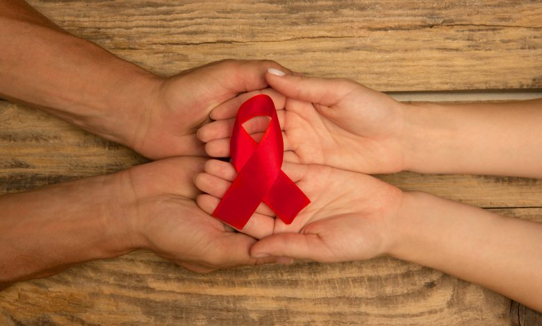 A imagem traz um laço vermelho, símbolo da conscientização do Dezembro Vermelho e do combate à pandemia da Aids, em mãos sobrepostas de duas pessoas.