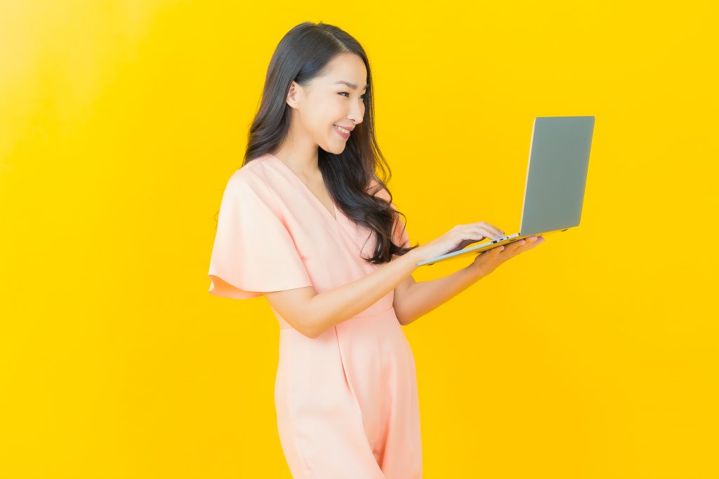 Mulher asiática sorridente de cabelo preto longo e macacão rosa claro em pé segurando um notebook cinza em um fundo amarelo.