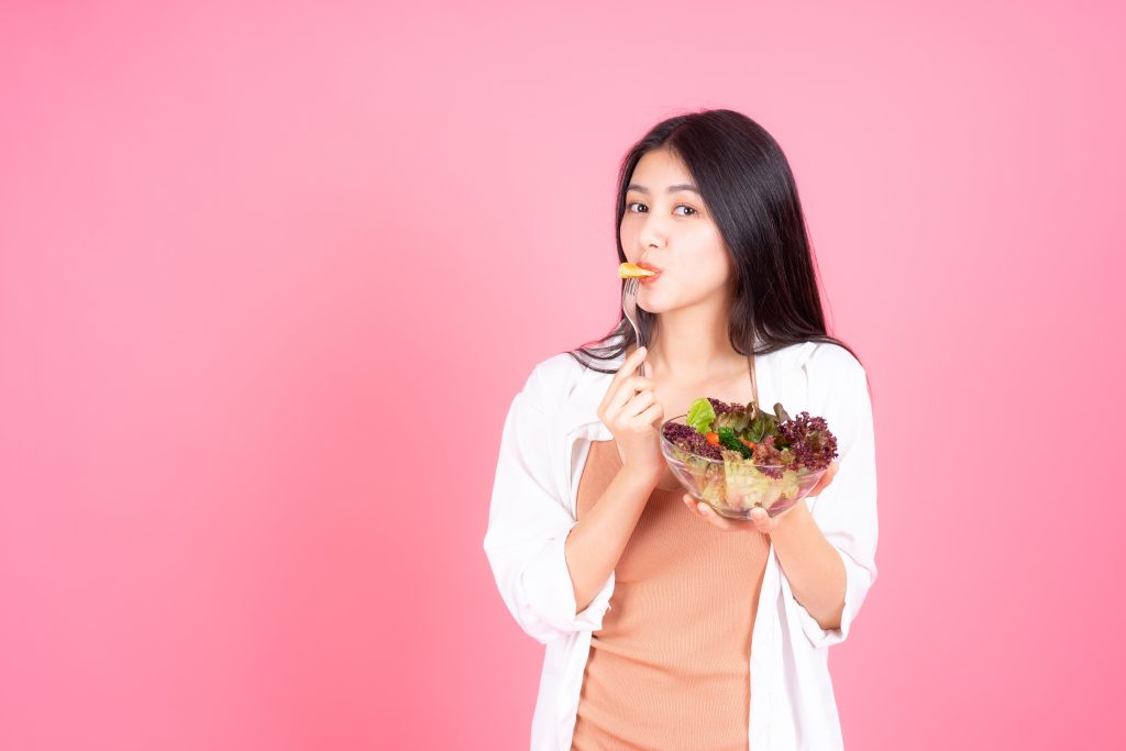 A imagem mostra uma mulher jovem e amarela, de cabelos pretos, comendo salada. Ela veste camiseta marrom, a qual está sobreposta por um blazer branco. Na mão direita, ela segura um pote de vidro com diversos legumes e verduras, enquanto, com a mão esquerda, ela leva um garfo à boca. Ao fundo, uma parede rosa.
