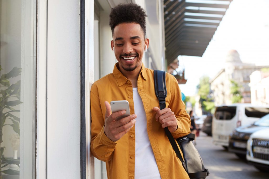 Um jovem negro de camisa amarela está escorado no muro de um prédio na rua enquanto sorri segurando seu smartphone.