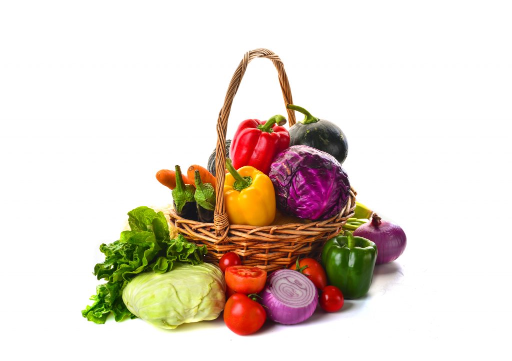 Vários legumes e frutas coloridos dentro e ao redor de uma cesta marrom com um fundo branco.
