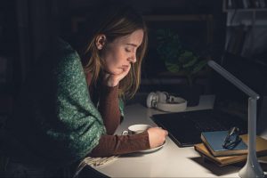 Uma mulher branca jovem encara uma xícara em cima de uma mesa com livros e um notebook em cima. É noite e a mulher tem um olhar cabisbaixo e exausto.