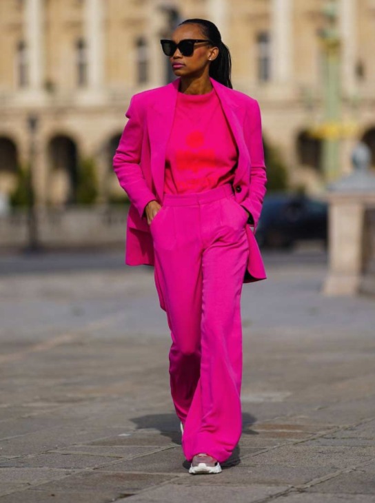Mulher anadando na rua com roupa monocromática rosa: calça, camiseta e blazzer. Usa um óculos de sol e está com as duas mãos no bolso
