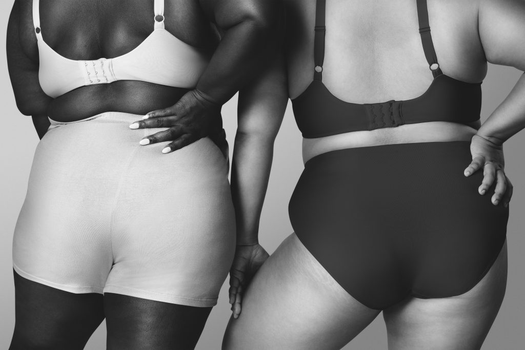 A imagem apresenta uma tonalidade branco e preto, com duas mulheres plus size de costas. Elas estão usando sutiã e calcinha short com a mão na cintura. A da esquerda é negra e a da direita é branca.