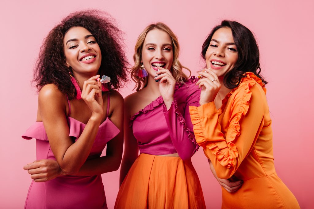 Três mulheres, a primeira negra, a segunda branca e loira e a terceira branca e morena. Estão usando roupas rosas e laranjas posando em um fundo rosa.