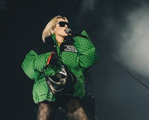 A cantora Miley Cyrus está em fundo preto com fumaça branca segurando um microfone e cantando. Está de óculos escuros, uma jaqueta puffer verde e segura uma bolsa preta, usa shorts preto e meia calça.