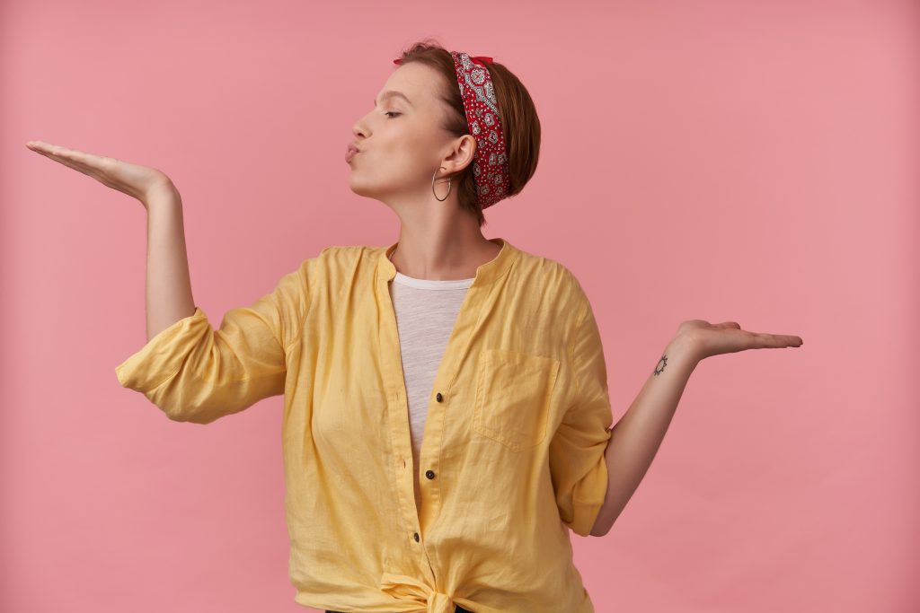 Mulher usando uma blusa branca por baixo de uma camiseta amarela desabotoada de manga longa, com uma bandana vermelha na cabeça, atrás de um fundo rosa claro. Está virada de lado e fazendo um bico com a boca com cada braço dobrado e direcionado para um lado
