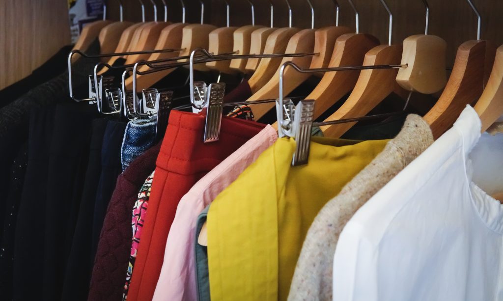 A imagem apresenta roupas em cabides pendurados em um armário, sendo elas nas cores preta, vermelha, rosa, amarela, branca e jeans.
