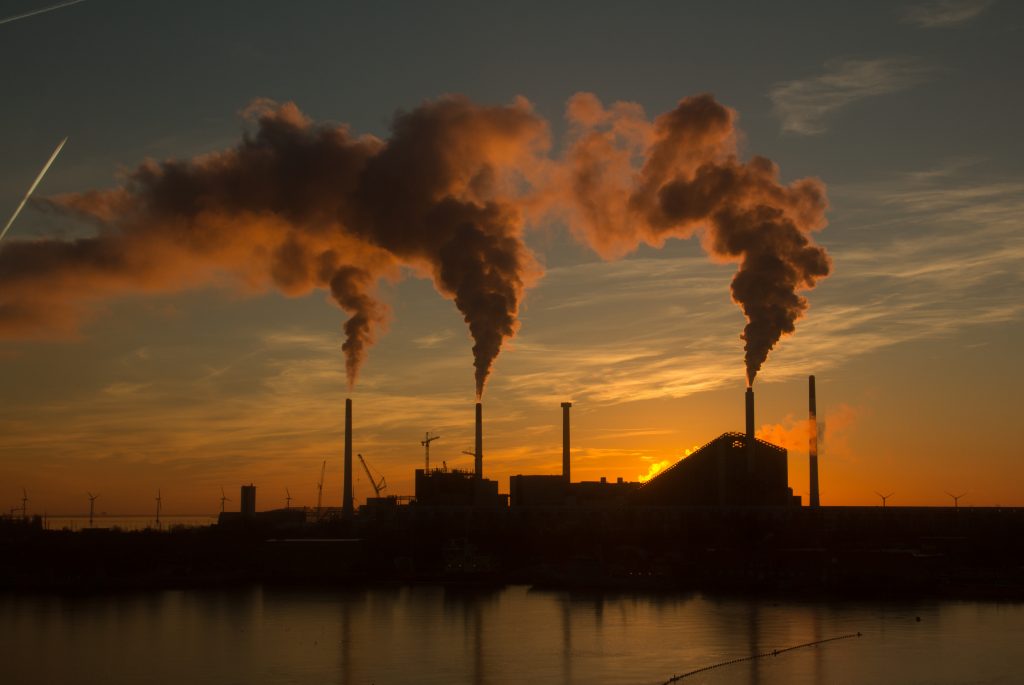 A imagem mostra uma fábrica soltando fumaça branca pelas chaminés, ao fundo podemos observar uma paisagem, o sol está se pondo atrás da construção.