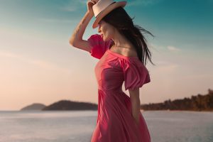 A imagem apresenta paisagem praiana, há uma mulher branca com cabelos pretos que está de lado, ela usa um vestido rosa. A Mulher utiliza um chapéu e segura-o com uma das mãos.