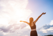 A imagem apresenta uma mulher branca de costas, estendo seus braços em direção ao céu, azul, com algumas nuvens e luz solar. A mulher tem cabelos castanhos e longos presos em um rabo de cavalo, enquanto veste top e calças pretos.