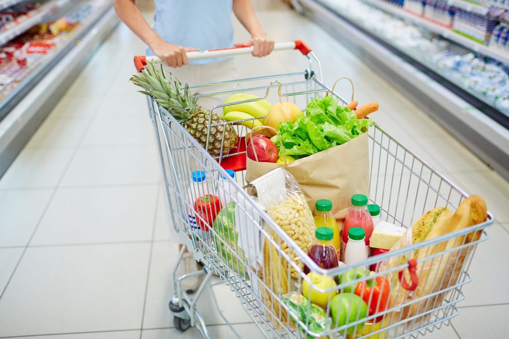 Mulher dentro de um supermercado segurando carrinho de compras cheio de sacolas com frutas e vegetais, pães e garrafas de suco.