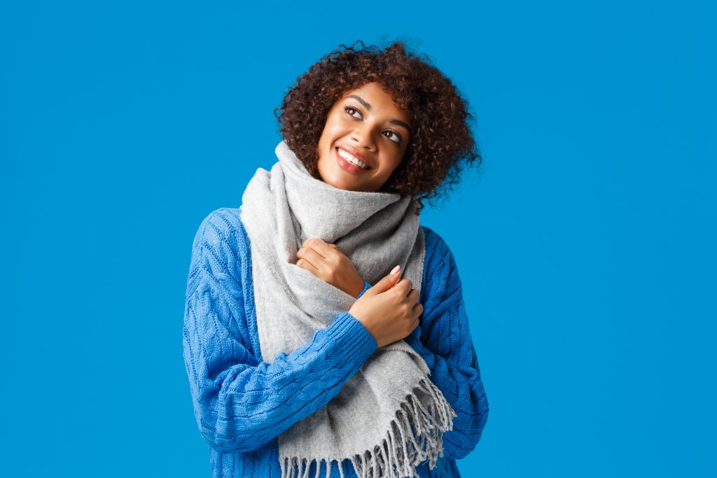 Mulher negra em um fundo azul sorri com as mãos cruzadas no peito em sinal de frio. Usa uma blusa azul e cachecol cinza