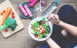 A imagem mostra diversos elementos relacionados a uma vida saudável: um prato de frutas, garrafas de água, cordas de exercício e alguns vegetais.