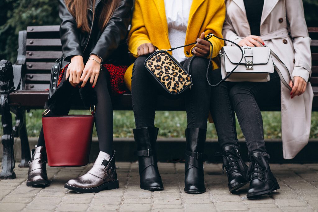 três mulheres sentadas em um banco com bolsas diferentes, só aparecem as pernas e as mãos delas