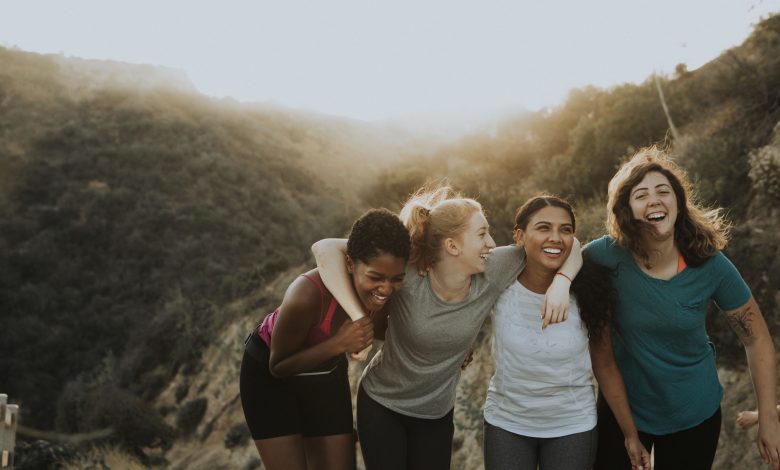 A imagem apresenta quatro amigas abraçadas e sorrindo com roupas fitness. O cenário ao fundo apresenta uma montanha.