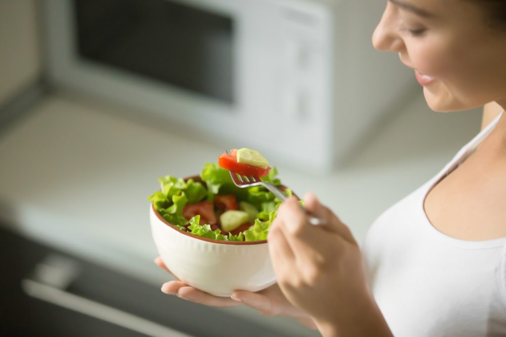 Mulher branca, em segundo plano, segurando uma tigela branca, em destaque, com salada de alface e tomate. Ela pratica o comer intuitivo, sustentando uma dieta saudável.