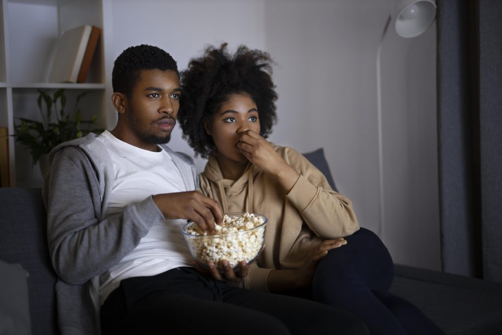 Casal comendo pipoca, de luz apagada, enquanto assiste um programa de TV 