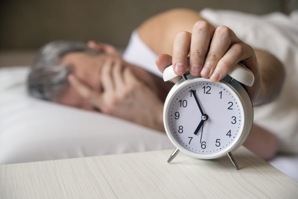  A imagem mostra um despertador enquanto ao fundo um homem de cabelos brancos está com a mão no rosto enquanto aberta para o despertador desligar.
