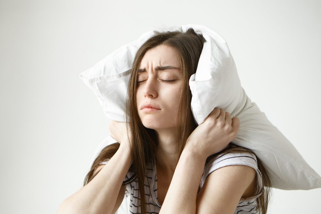 Imagem de uma mulher branca de cabelos castanhos segurando um travesseiro branco em cima da cabeça enquanto está de olhos fechados e sobrancelha franzida. 