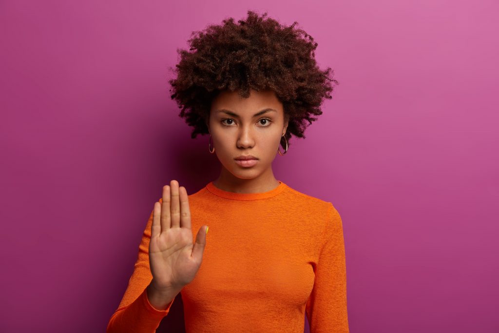 Mulher negra com a mão esquerda levantada vestindo blusa de manga longa laranja em um fundo roxo. Ela está aprendendo a dizer não, uma prática necessária e que pode ser aprendida.