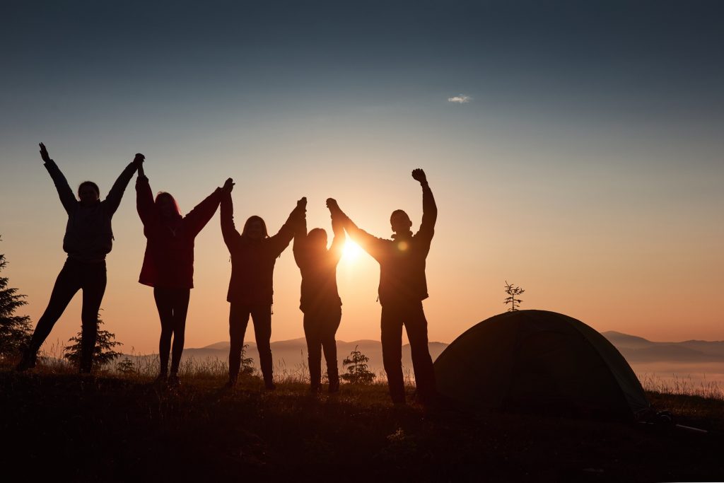 Foto que mostra a silhueta de 5 pessoas com as mãos dadas observando o pôr do sol. A paisagem natural é composta por montanhas e árvores. A imagem mostra benefícios de dizer “não”.
