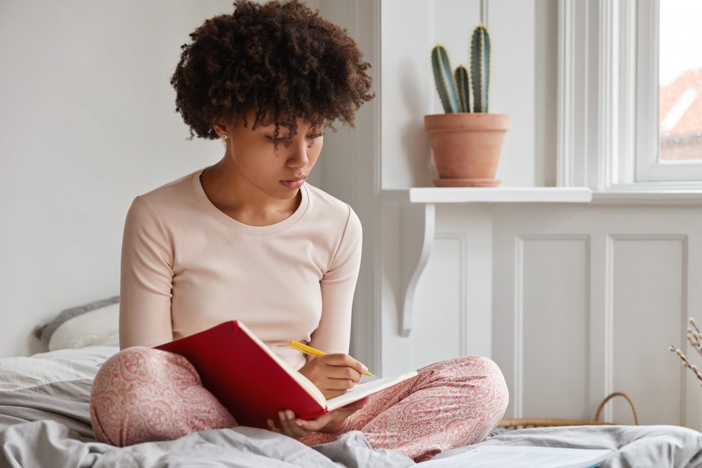 mulher negra usando um pijama rosa claro está sentada na cama com as pernas cruzadas escrevendo em seu diário