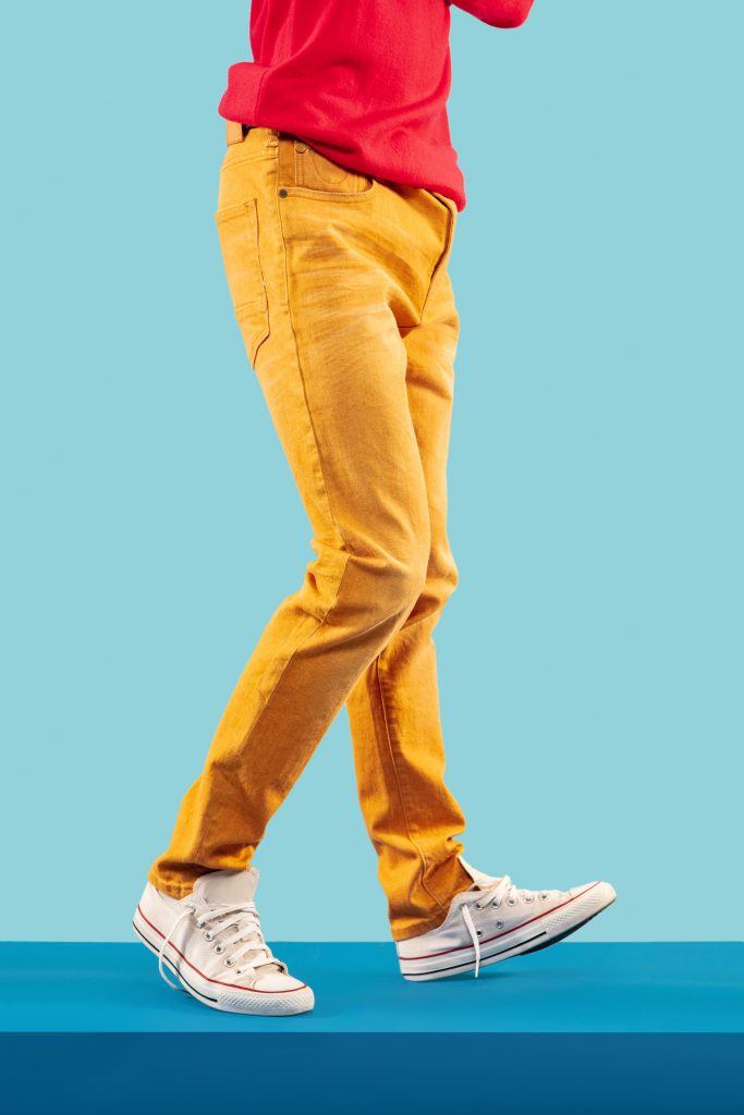 Metade do corpo de homem em um fundo azul usando camiseta vermelha, calça amarela e all star branco