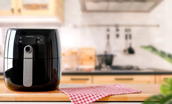 airfryer de cor preta em um balcão de cozinha de madeira. Ao lado, há um pano de prato quadriculado vermelho e branco. O restante da cozinha aparece ao fundo, mas desfocada