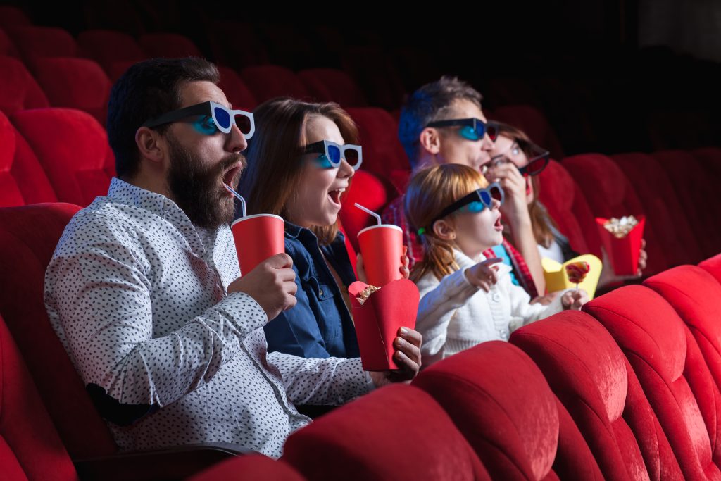 O cenário da foto é uma sala de cinema com poltronas vermelhas. Na fileira central, há um grupo de quatro adultos e uma criança assistindo um filme. Todos usam óculos 3D e seguram copos vermelhos e embalagens de pipocas na mão. 